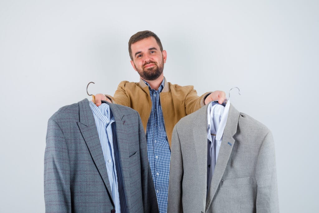 A man holding two different suits, a gray one and a checked one, in front of him while smiling and looking at the camera.