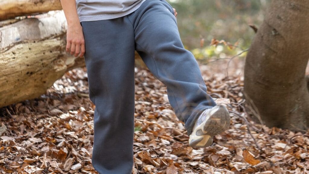 "Close-up of a person wearing gray sweatpants and sneakers, walking through a forest floor covered with dry leaves. The focus is on their legs as they take a step, with a tree and nature background."
