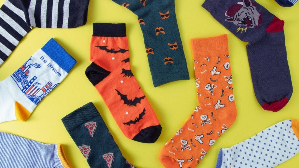 A flat lay of colorful Halloween-themed socks on a bright yellow background, featuring patterns like bats, skulls, pumpkins, stripes, and cityscape designs