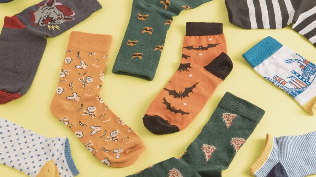 A flat lay of colorful socks laid out on a pale yellow background. The socks feature various patterns: an orange sock with small skull and bone prints, a green sock with bats, an orange sock with black toes and heel, a dark green sock with pizza slice motifs, a white sock with blue trim and a cityscape scene, a light blue polka-dotted sock, and other striped or patterned socks partially visible at the edges. The arrangement is scattered and playful.