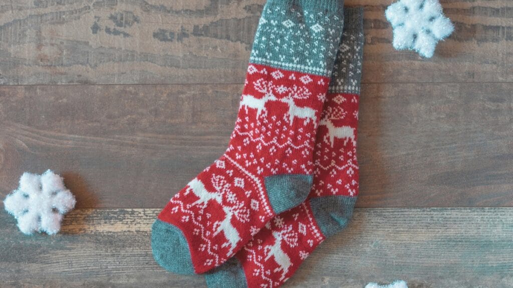 A pair of knitted winter socks with a red and white reindeer and snowflake pattern, paired with teal accents at the toes, heels, and cuffs. The socks are laid diagonally on a rustic wooden surface, with two light blue artificial snowflakes nearby for a festive touch.