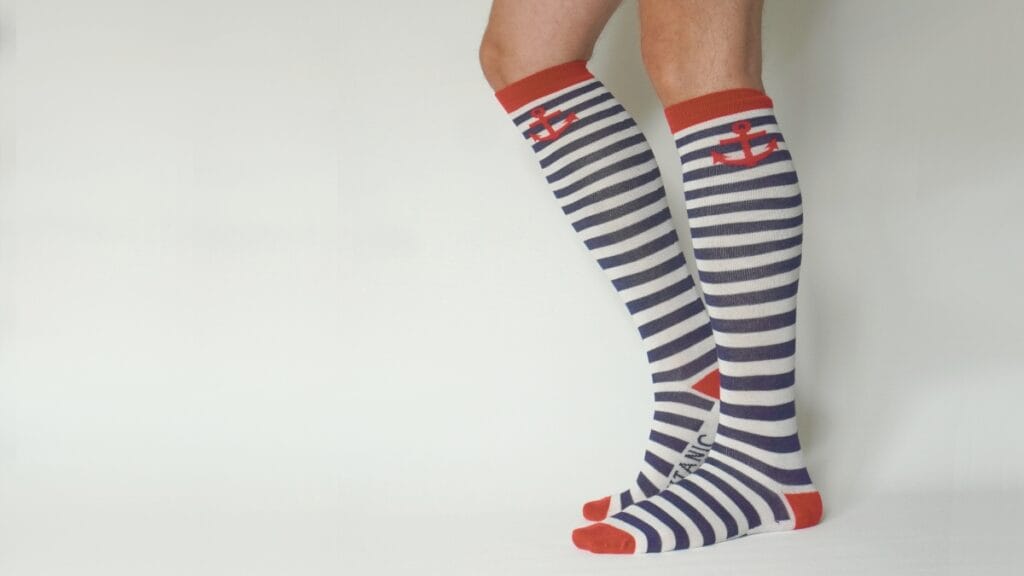 A pair of legs wearing navy and white horizontal striped knee-high socks with red accents. The socks feature red toes, heels, and top bands, and display a red anchor motif near the top. The person is standing on a light-colored background.