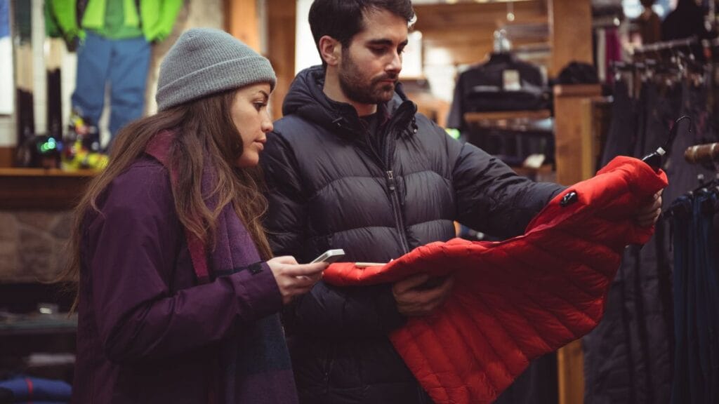 A man and woman are shopping for winter jackets in a store. The man is holding a red puffer jacket, while the woman looks at her phone.