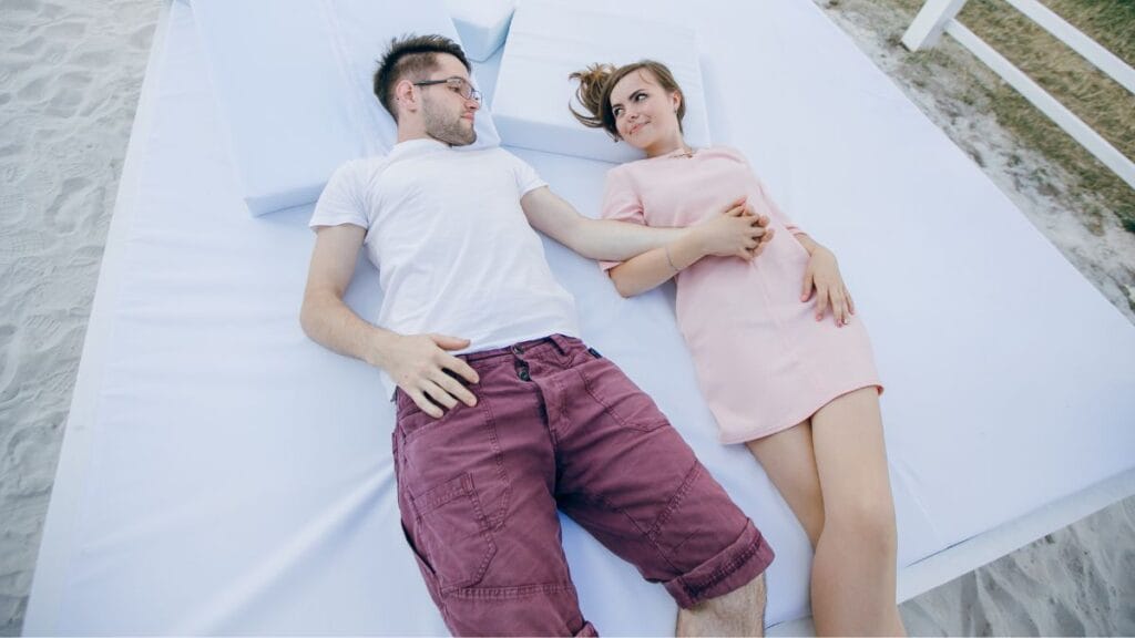  A couple holding hands, wearing casual outfits—one in a white blouse and blue jeans, the other in a grey t-shirt and olive green shorts—standing by the water.