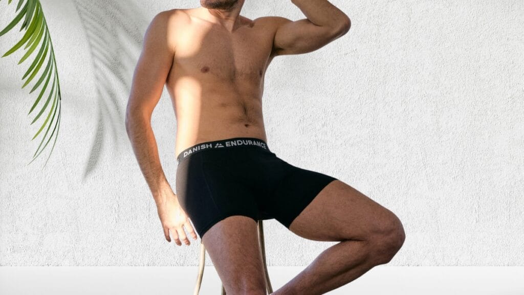 A shirtless man wearing black boxer briefs with a white waistband that reads ‘DANISH ENDURANCE’ around the waist, posing against a light textured wall.