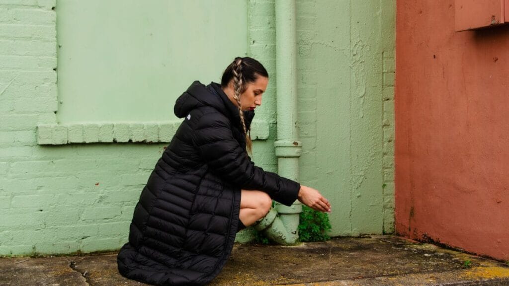 A woman wearing a black puffer jacket crouches down beside a green wall, examining something on the ground.