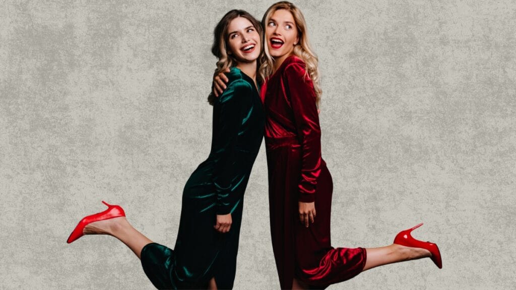 Two women posing in red and green velvet fall dresses, smiling with red heels