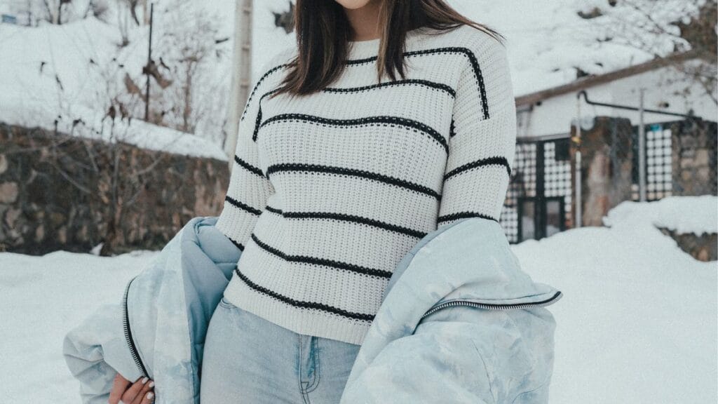 A woman wearing a white striped sweater and light blue jacket standing outside in the snow.