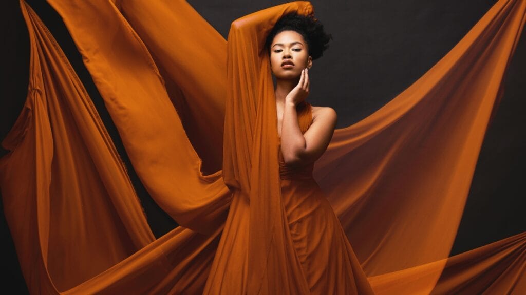 Model wearing an orange dress made of sheer fabric, flowing beautifully in a dramatic pose