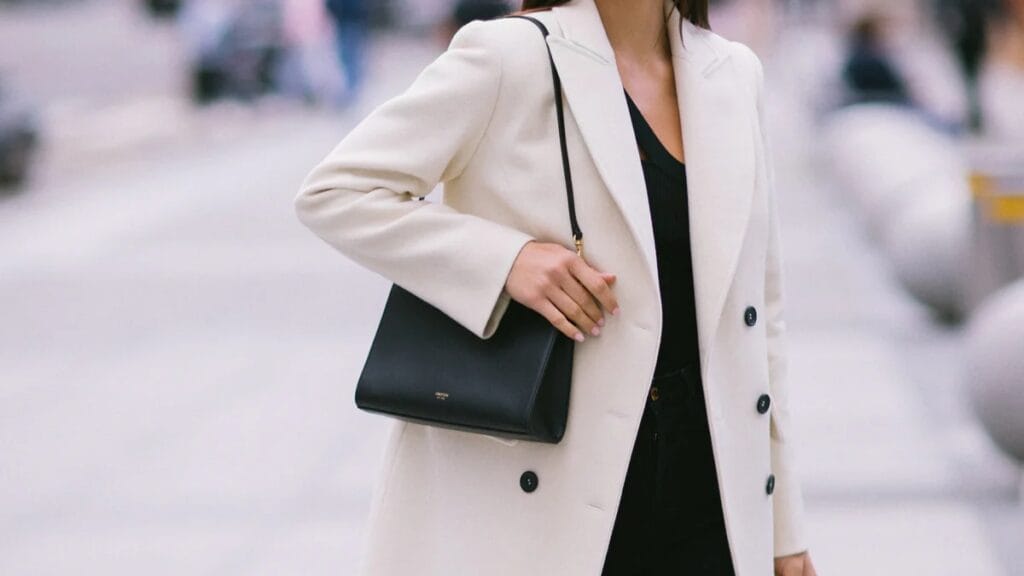 A woman wearing a cream-colored double-breasted coat over a black top stands outdoors. She carries a small black rectangular shoulder bag with a thin strap. The background is blurred, suggesting a city street.