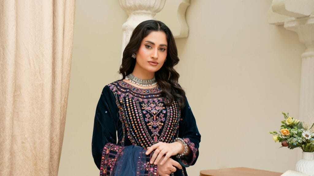 Elegant woman standing indoors wearing a dark embroidered traditional outfit with colorful patterns, a jeweled necklace, and bangles, posed with one hand resting near her waist.