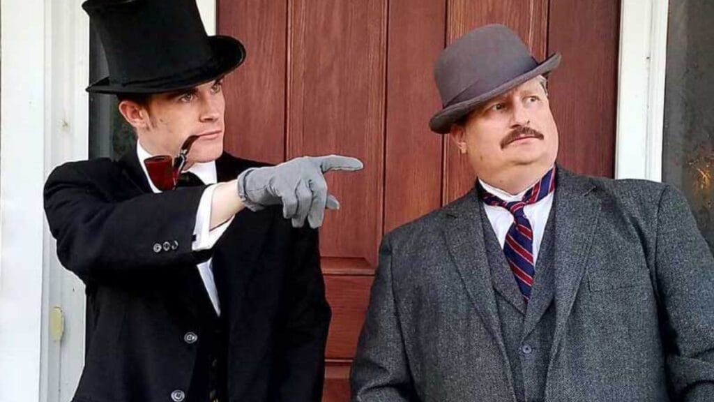 A young man in 19th-century French-inspired clothing, wearing a dark overcoat, black top hat, and cravat, standing indoors with another suited man behind him.