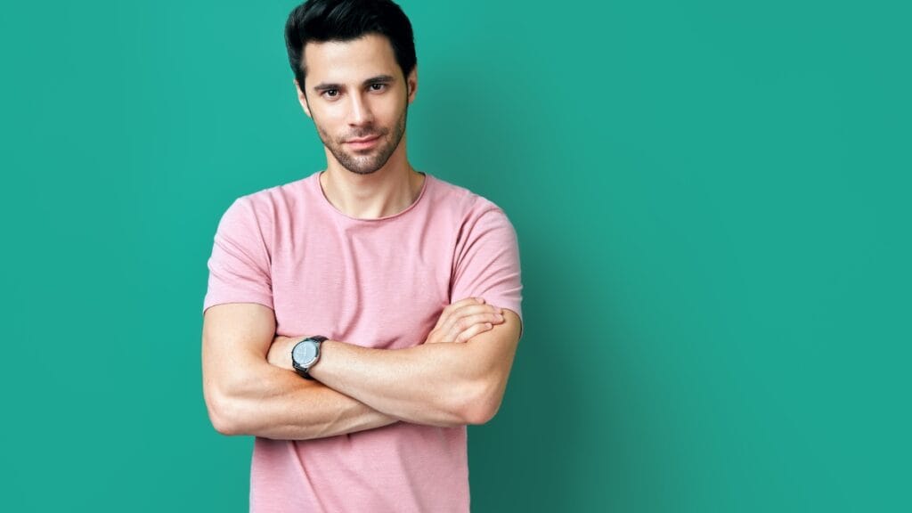 A man in a pink short-sleeve shirt standing with his arms crossed, wearing a wristwatch, against a solid teal background.