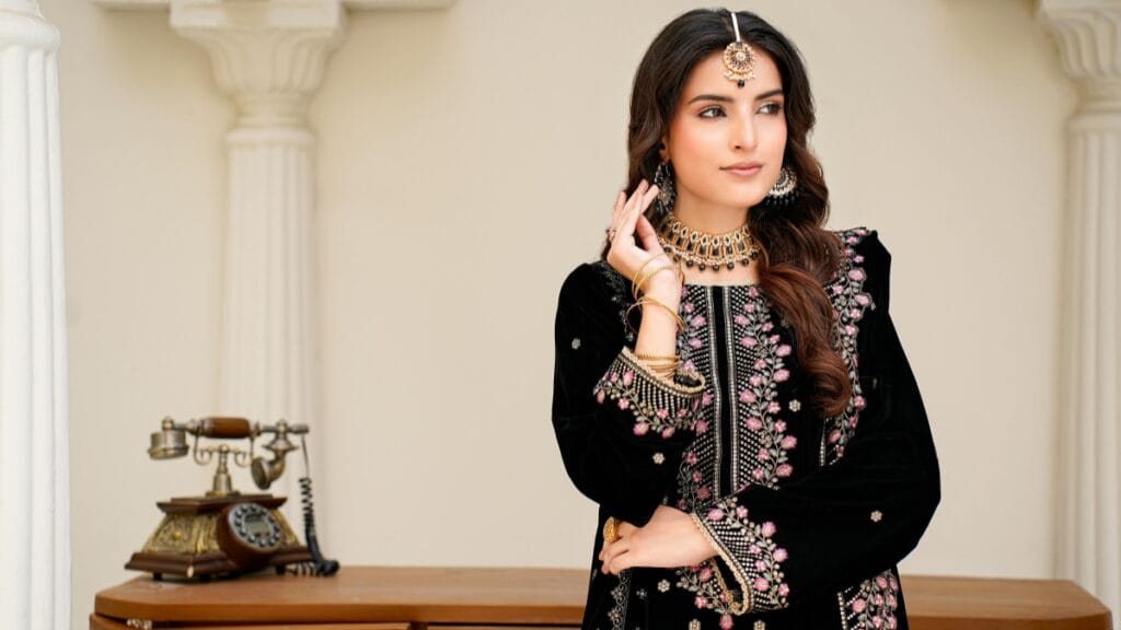 Elegant woman in a black embroidered traditional outfit with pink floral detailing, layered gold jewelry, and a maang tikka, posing indoors beside a vintage telephone.