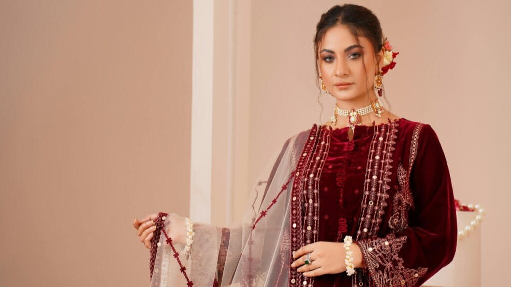 Elegant woman in a deep burgundy traditional outfit with intricate embroidery and layered jewelry, standing indoors and holding a sheer dupatta.