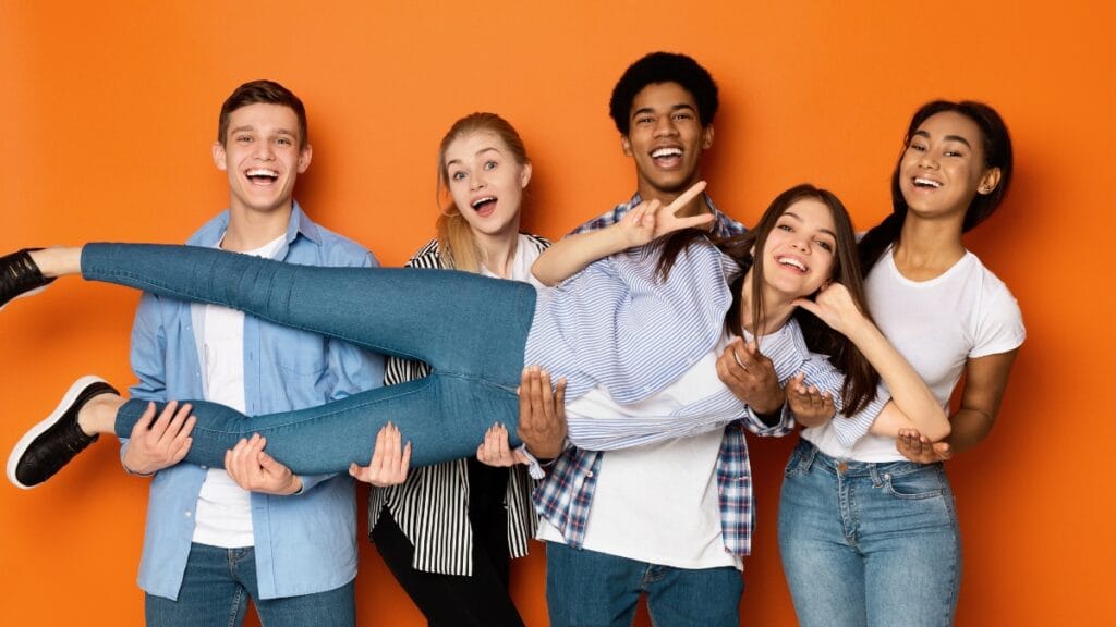 A group of five young people pose and laugh together in front of an orange background, wearing denim jeans and casual tops.