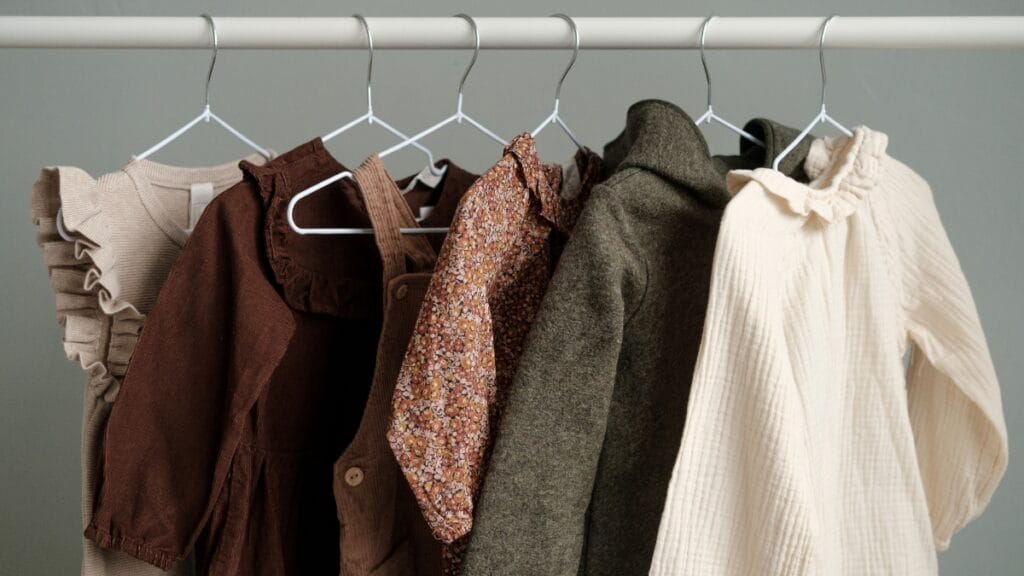 Close-up of hanging clothing on white hangers: brown jackets, a brown patterned shirt, a gray coat, and a cream ruffled knit sweater, displayed against a light gray background.