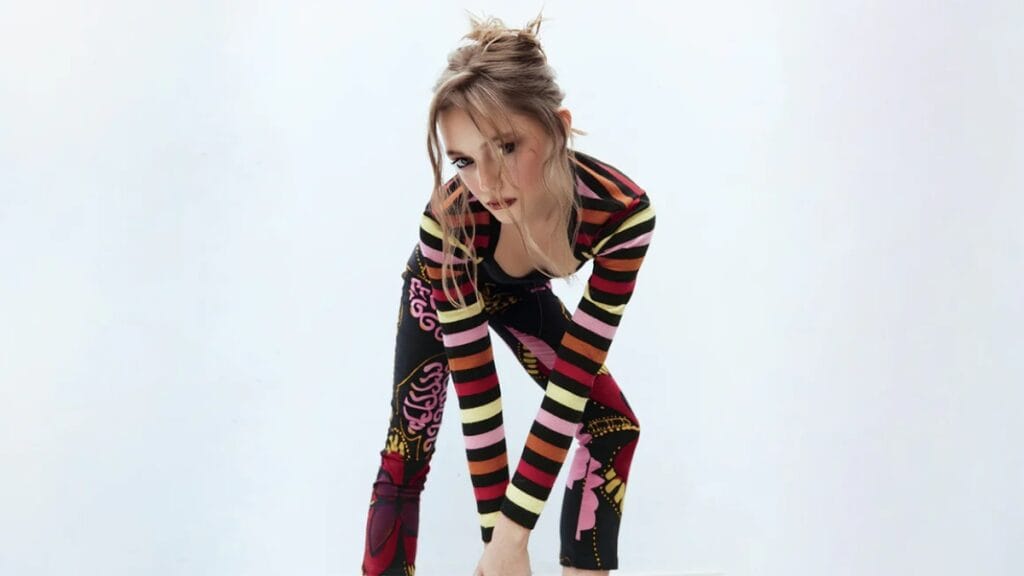 A model poses in a striped black, pink, and yellow long-sleeve outfit during a fashion shoot, looking downward with hair styled up