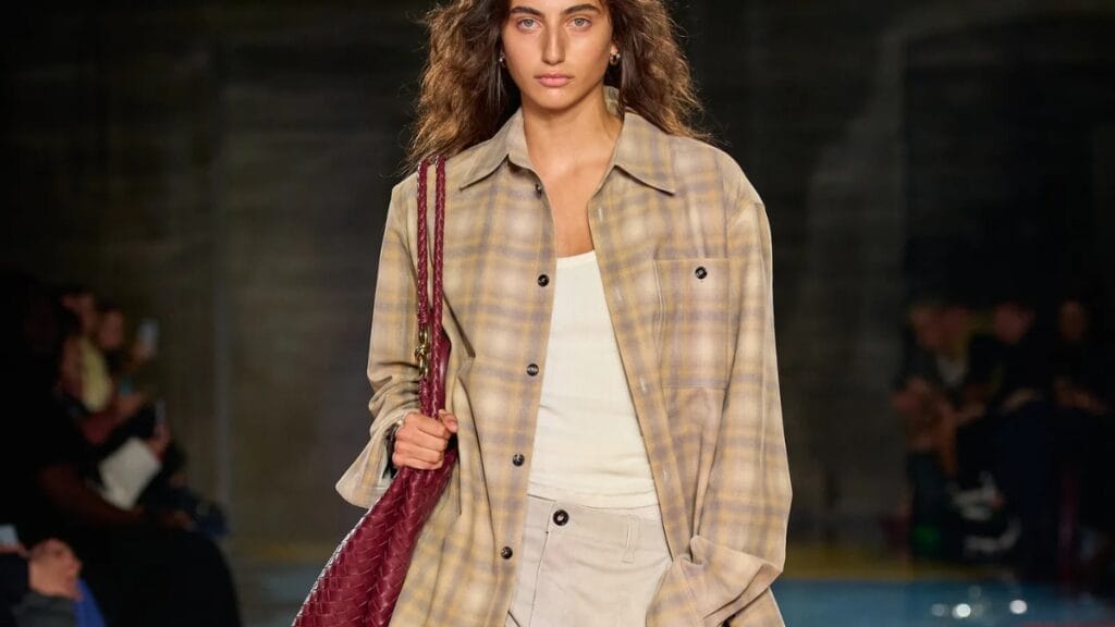 A model walks the runway wearing a light plaid overshirt over a white outfit, carrying a burgundy textured bag.