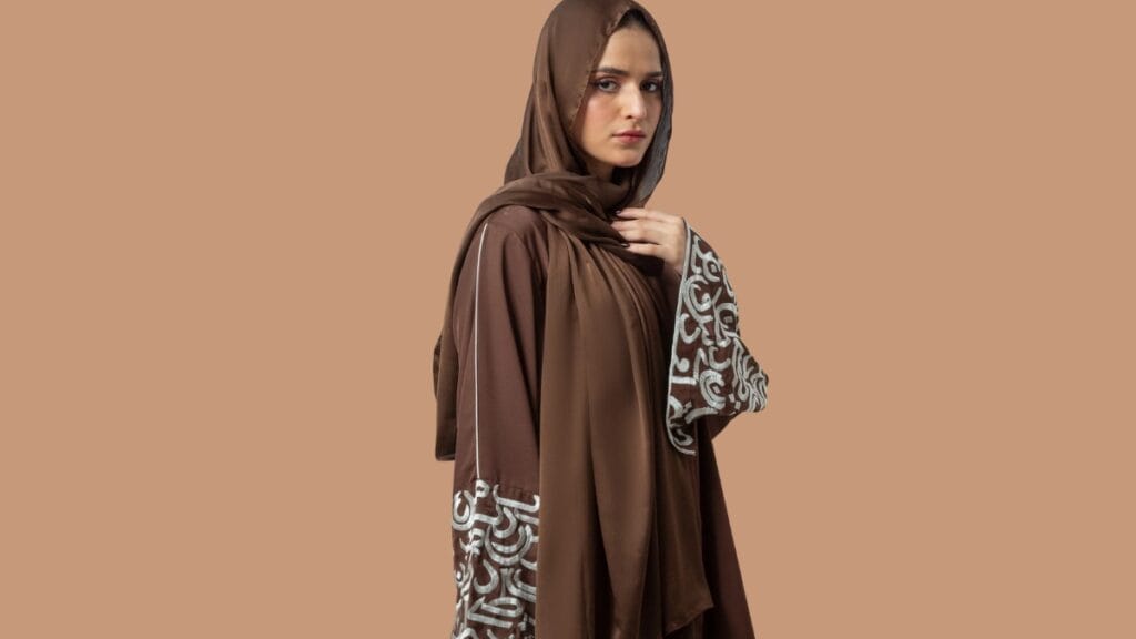 A woman wearing a brown abaya with intricate white embroidery on the sleeves, paired with a matching brown hijab, standing against a neutral background.