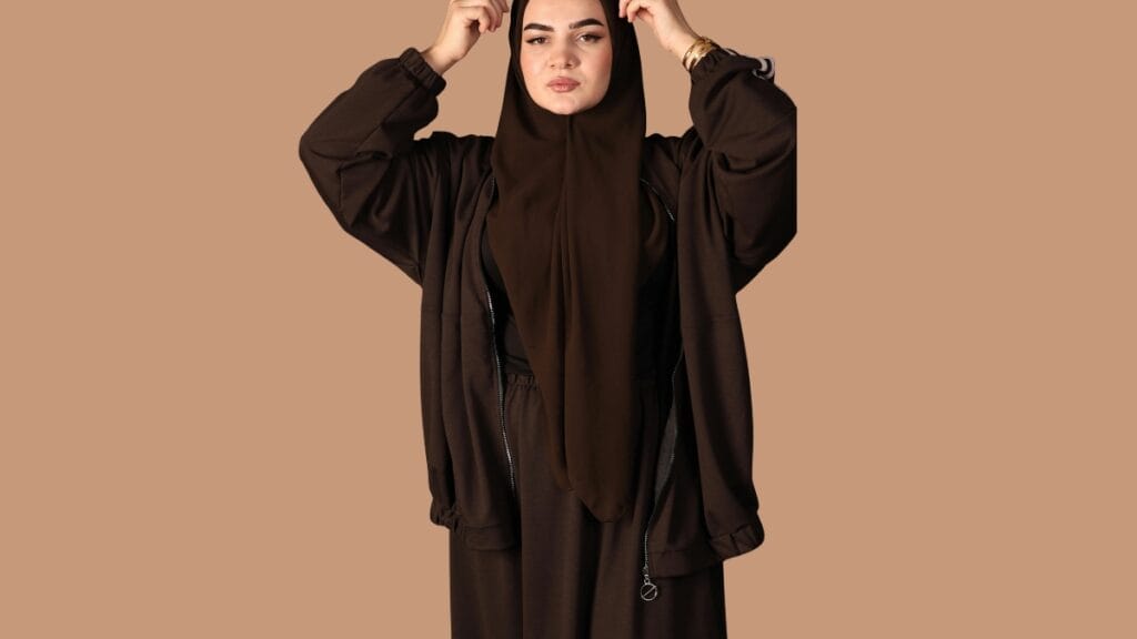 A woman in a brown abaya and beige hijab, holding a beige handbag, with sunglasses on, standing against a neutral background.