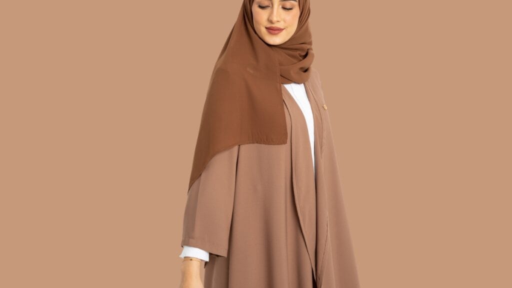 A woman wearing a beige abaya with a matching brown hijab, standing against a neutral background, exuding a graceful and modest style.