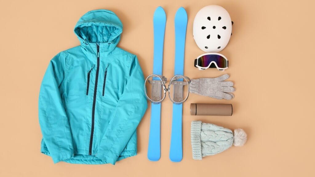 A flat-lay of winter sports gear—blue jacket and ski boots, ski goggles, foam skis, gloves, and a snowboard-style helmet—on a beige background.