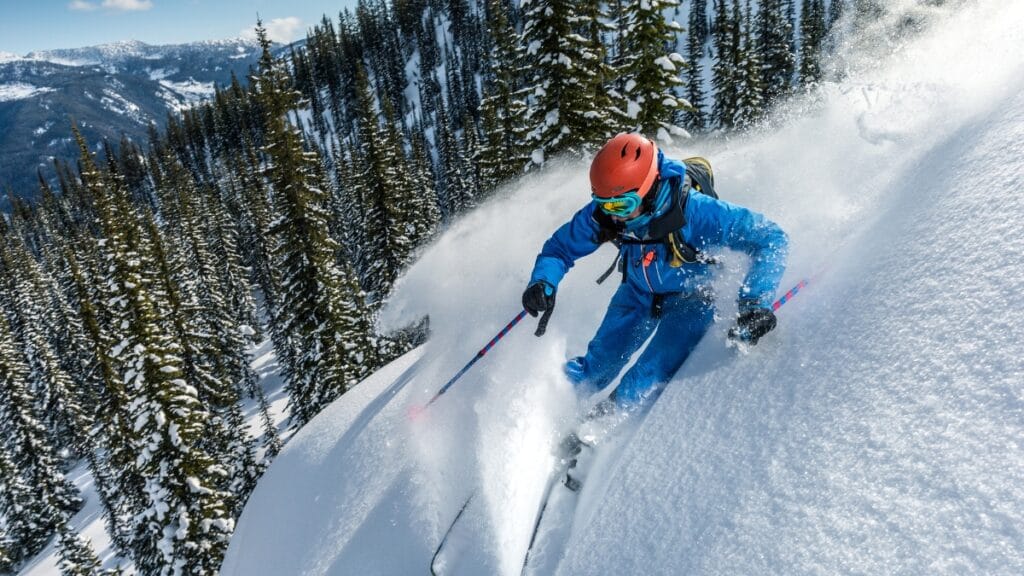 A skier in blue winter gear carves through fresh snow on a mountain slope, with pine trees lining the background and a spray of snow kicked up behind them.