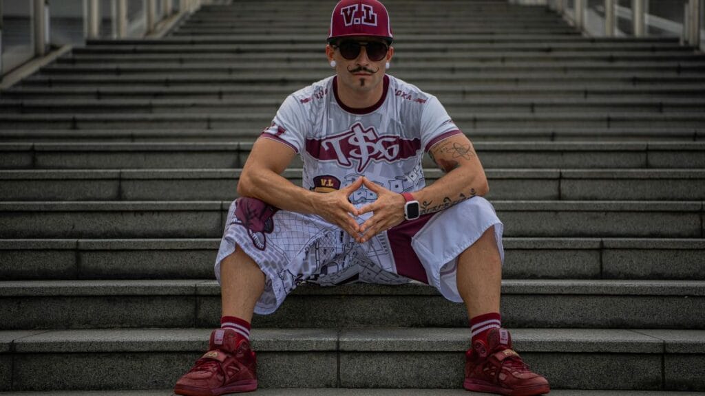 Man in streetwear outfit sitting on concrete stairs wearing a cap, sunglasses, graphic t-shirt, shorts, and red sneakers