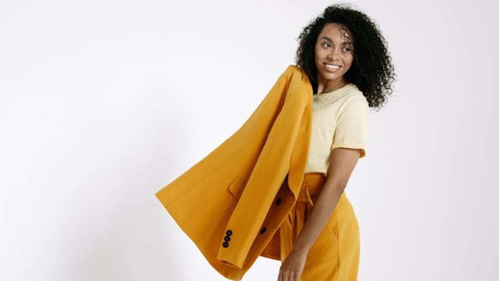 Smiling woman wearing a yellow outfit holding a matching blazer against a plain background