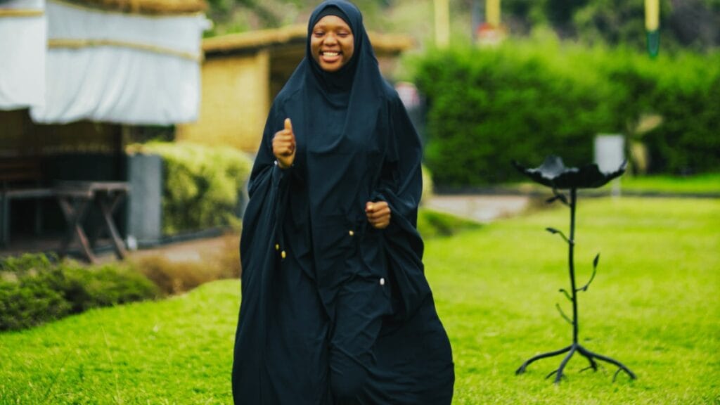 A person wearing a dark blue abaya stands outdoors on a green lawn, smiling and giving a thumbs-up with one hand; a black metal stand is visible to the right in the garden background.