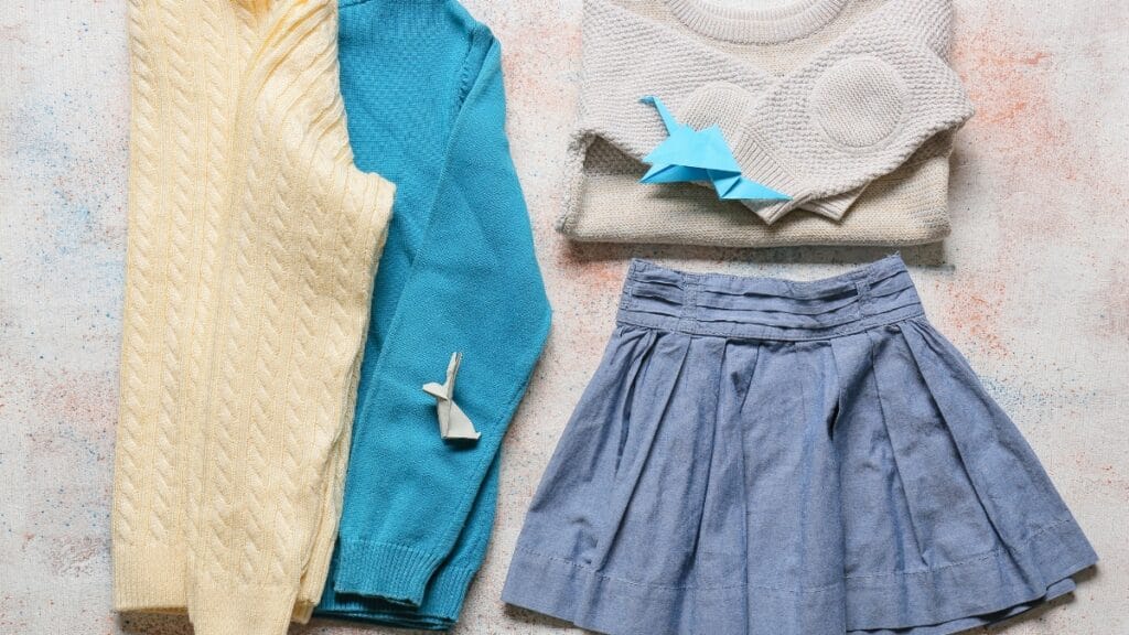 A flat lay of a cream cable-knit sweater, a teal sweater, a folded white knit top, a gray pleated skirt, and a few folded paper items on a light textured background.