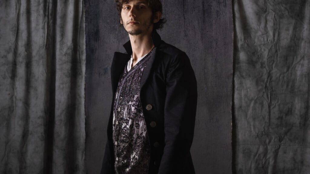 A young man wearing a black double-breasted jacket layered over a patterned medieval-style shirt, standing in front of a dark gray draped background.