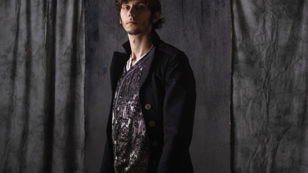 A young man wearing a black double-breasted jacket layered over a patterned medieval-style shirt, standing in front of a dark gray draped background.