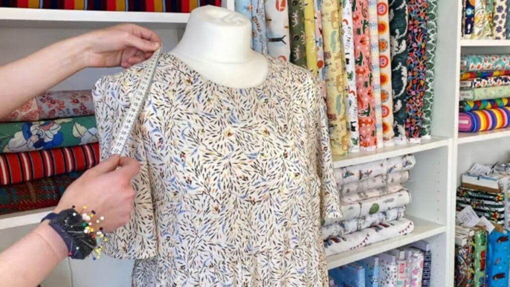 Measuring a patterned dress on a mannequin in a fabric shop with colorful textiles in the background.