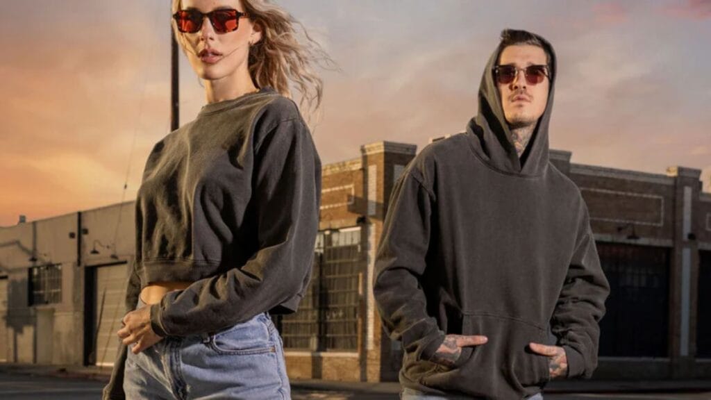 Two models wearing casual streetwear, including hoodies and sunglasses, standing outdoors in an urban setting at sunset.