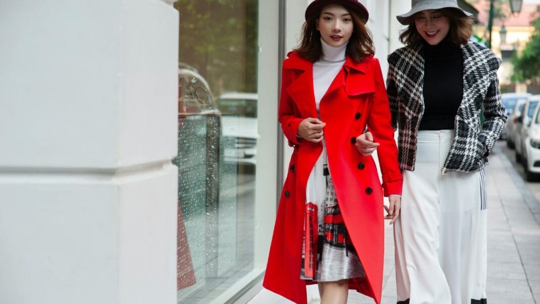 Two women walking on a city street wearing stylish coats, one in a bright red trench coat and the other in a patterned jacket with a hat.
