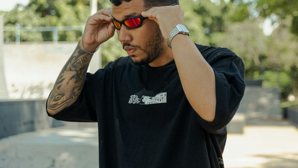 Man wearing a black streetwear t-shirt adjusting reflective sunglasses outdoors with a casual urban style look