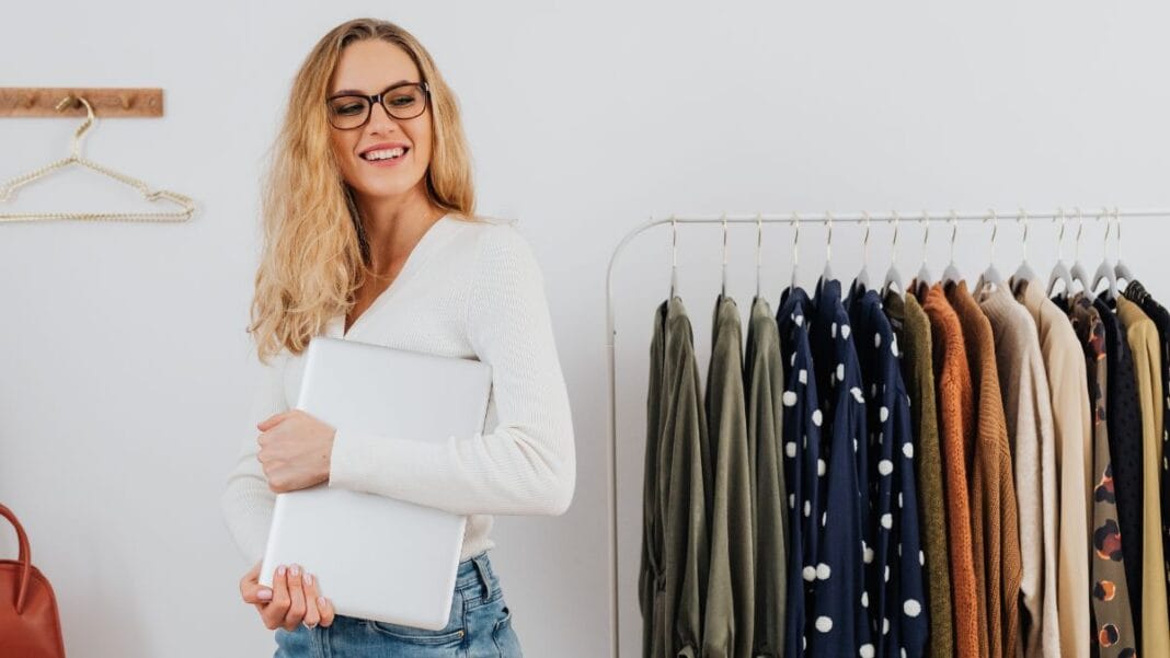 Woman holding a laptop standing beside a clothing rack with neatly arranged garments in a fashion workspace