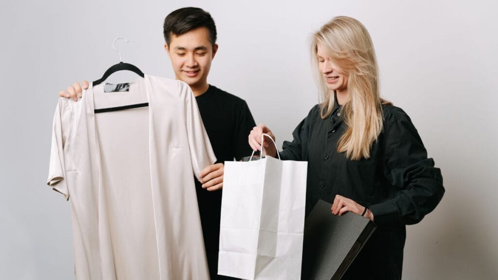Two fashion professionals reviewing a garment, one holding a dress on a hanger while the other checks a shopping bag in a studio setting