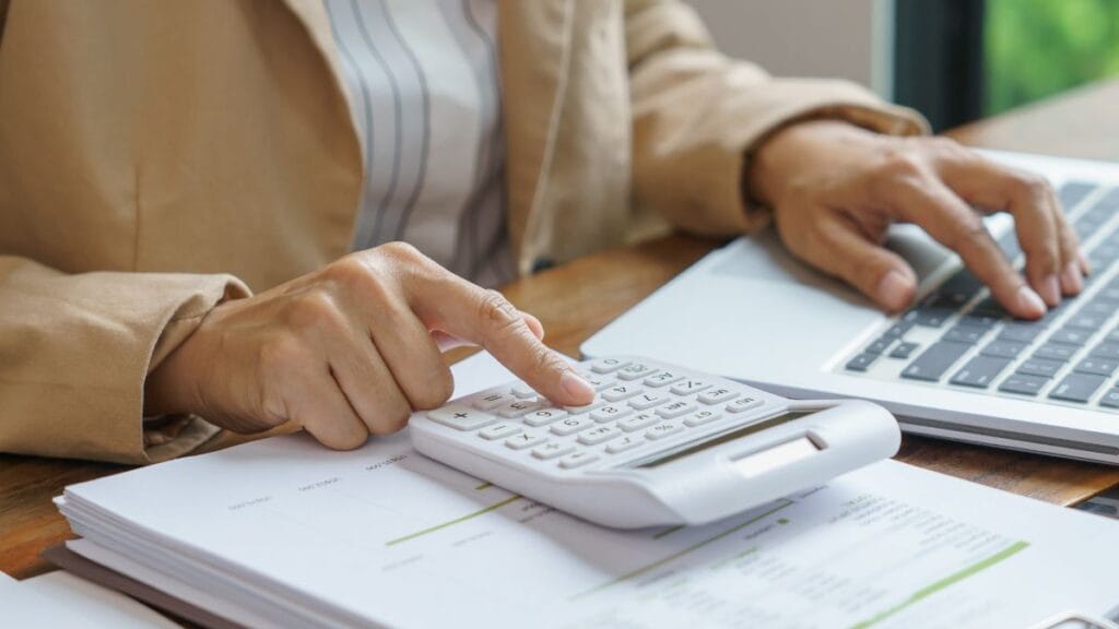 Person using a calculator and laptop to review financial documents and calculate costs.
