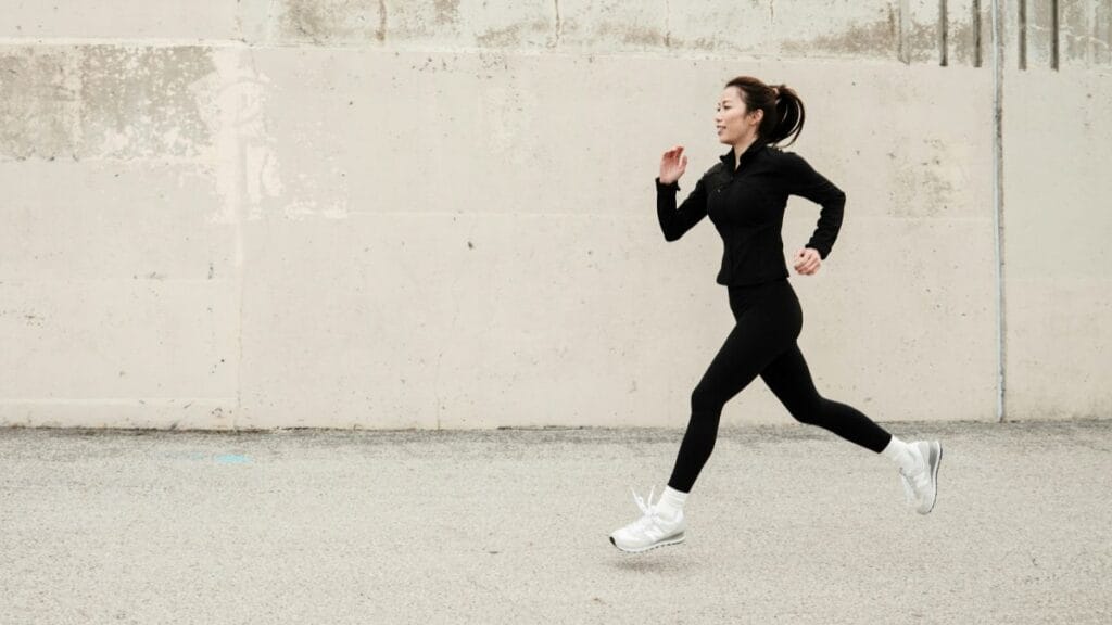Woman in black athletic outfit running along a concrete wall outdoors, wearing white sneakers.