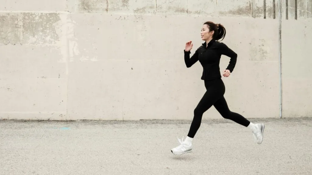 Woman in black athletic outfit running along a concrete wall outdoors, wearing white sneakers.