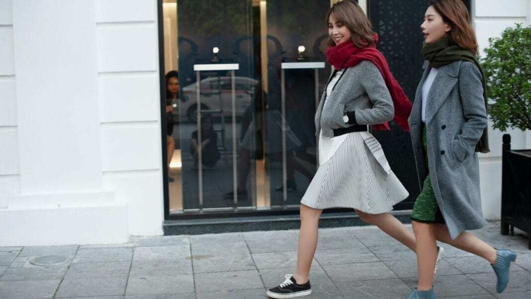 Two women in stylish coats and scarves walk side by side on a city sidewalk past a modern storefront.