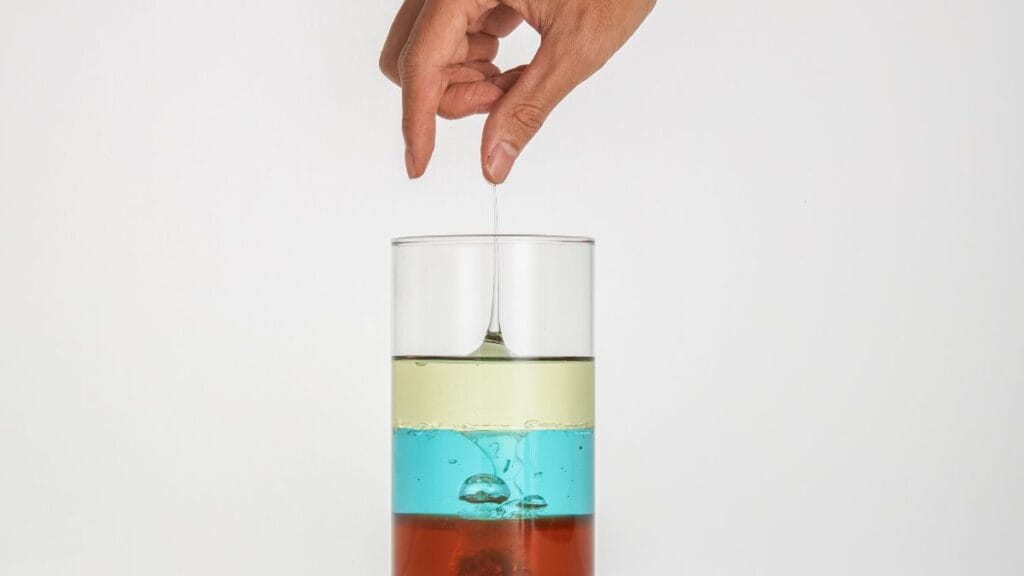 Hand pulling a thread of liquid from a layered glass, showing different viscosities and fluid behavior in a simple experiment