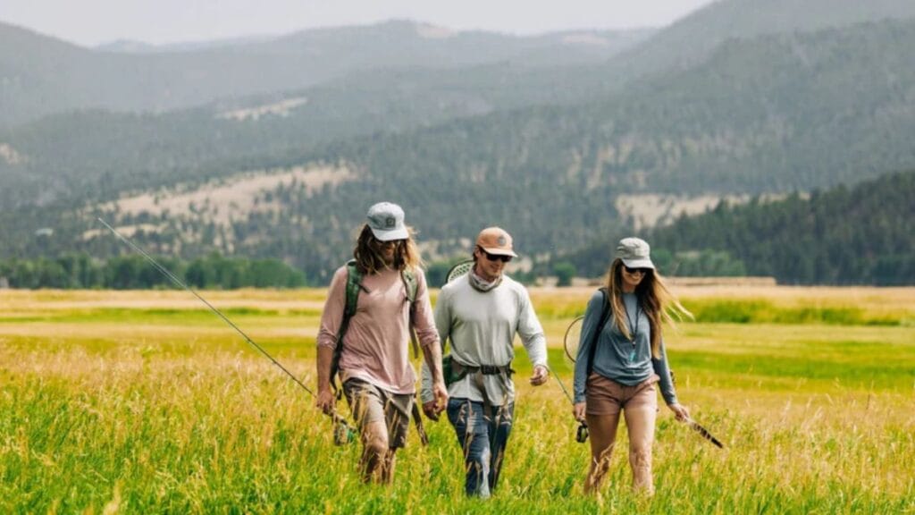 Three people walking through a grassy field with fishing rods in a scenic mountain landscape.