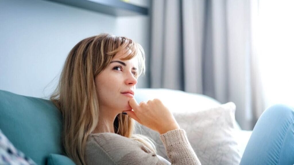 Woman sitting on a sofa looking thoughtful in a bright living room.