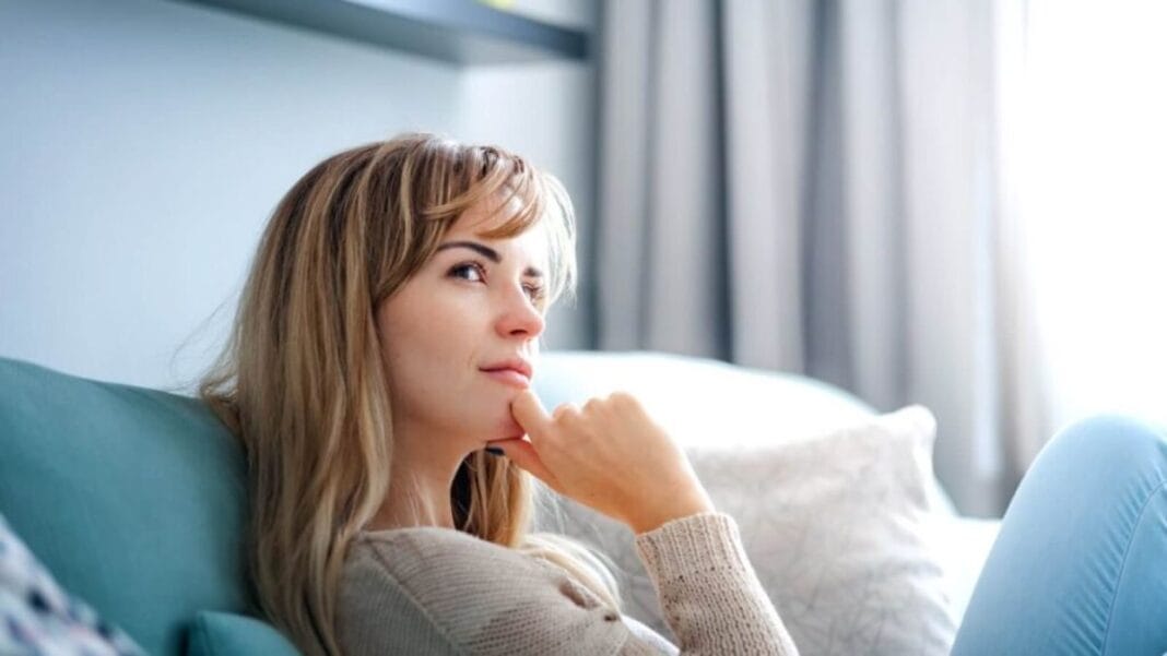 Woman sitting on a sofa looking thoughtful in a bright living room.