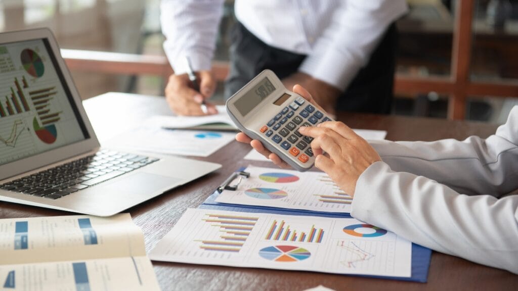 Write for Us Finance — professionals calculating figures with a calculator during a meeting with financial charts and documents.