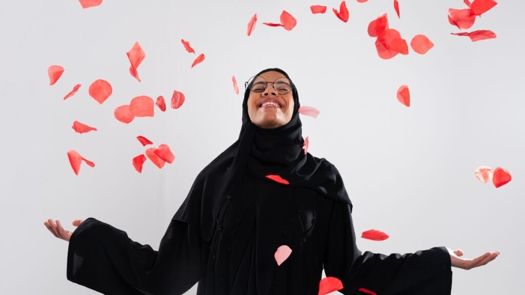 A joyful woman in a black abaya and hijab, smiling and surrounded by floating rose petals.